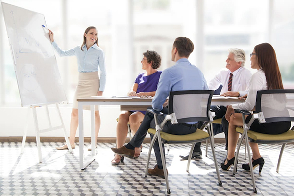 Woman stood at a whiteboard, training a group of employees that are sat at a desk.